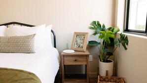 A neatly made bed with white linens and a patterned pillow, a bedside table with a framed picture and a small vase, a potted plant, and a wicker basket. - Vega 02: A spacious, 3-bedroom desert oasis stay, Vega #2 (Pinal County)