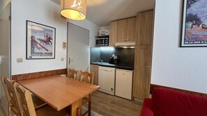 Cozinha compacta com armários de madeira, micro-ondas e máquina de lavar louça. Há uma mesa de jantar com cadeiras e um sofá vermelho na sala de estar.