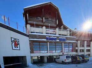 Un edificio con un letrero que dice "PLAGNE SOLEIL" y logotipos "esf". Hay vehículos estacionados frente a él, y el suelo está cubierto de nieve.