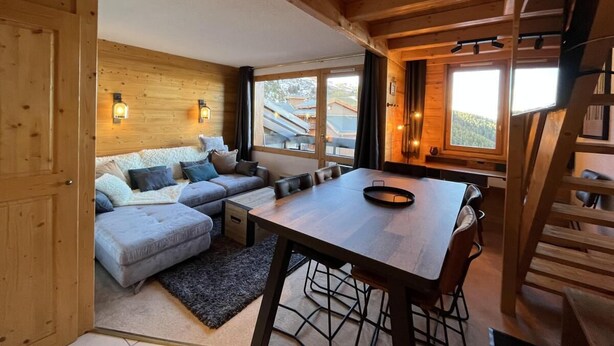 Un accogliente interno di una baita con soffitto in legno, un tavolo da pranzo con sedie, un divano con cuscini e una scala.