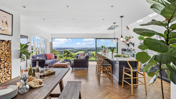 A modern living space with a large window overlooking a scenic landscape, a wooden dining table, and a kitchen area with a bar. - Fox Hill House in Haytor (Newton Abbot)