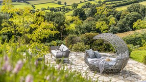 A garden patio with a wicker chair and a sofa, surrounded by lush greenery and a stone floor. - Fox Hill House in Haytor (Newton Abbot)