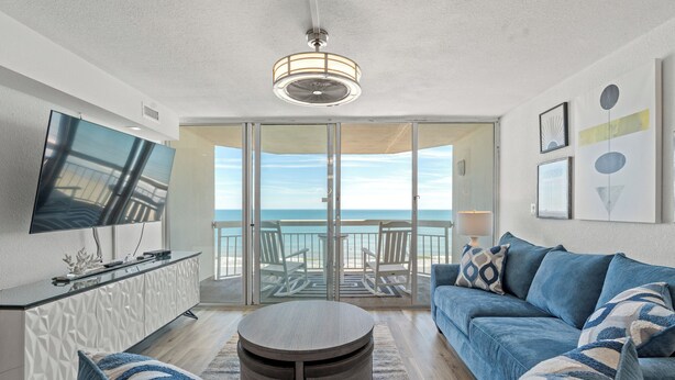 Un soggiorno moderno con una grande TV, un tavolino rotondo, un divano e un balcone con vista sulla spiaggia.