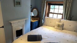 Een slaapkamer met een bed, kussens, een nachtkastje, een spiegel, een raam met gordijnen en een open haard.