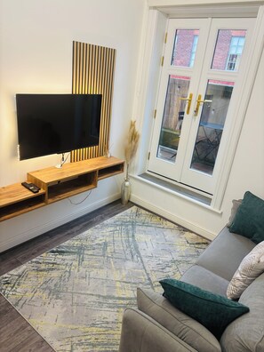 Ein modernes Wohnzimmer mit einem Flachbildfernseher an einer Holzwand, einem grauen Sofa mit grünen Kissen und einem gemusterten Teppich.