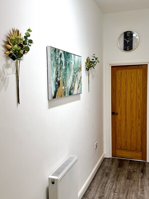 Ein Flur mit einem weißen Heizkörper, einer Holztür und Wandbildern, darunter eine Uhr und Blumenarrangements.