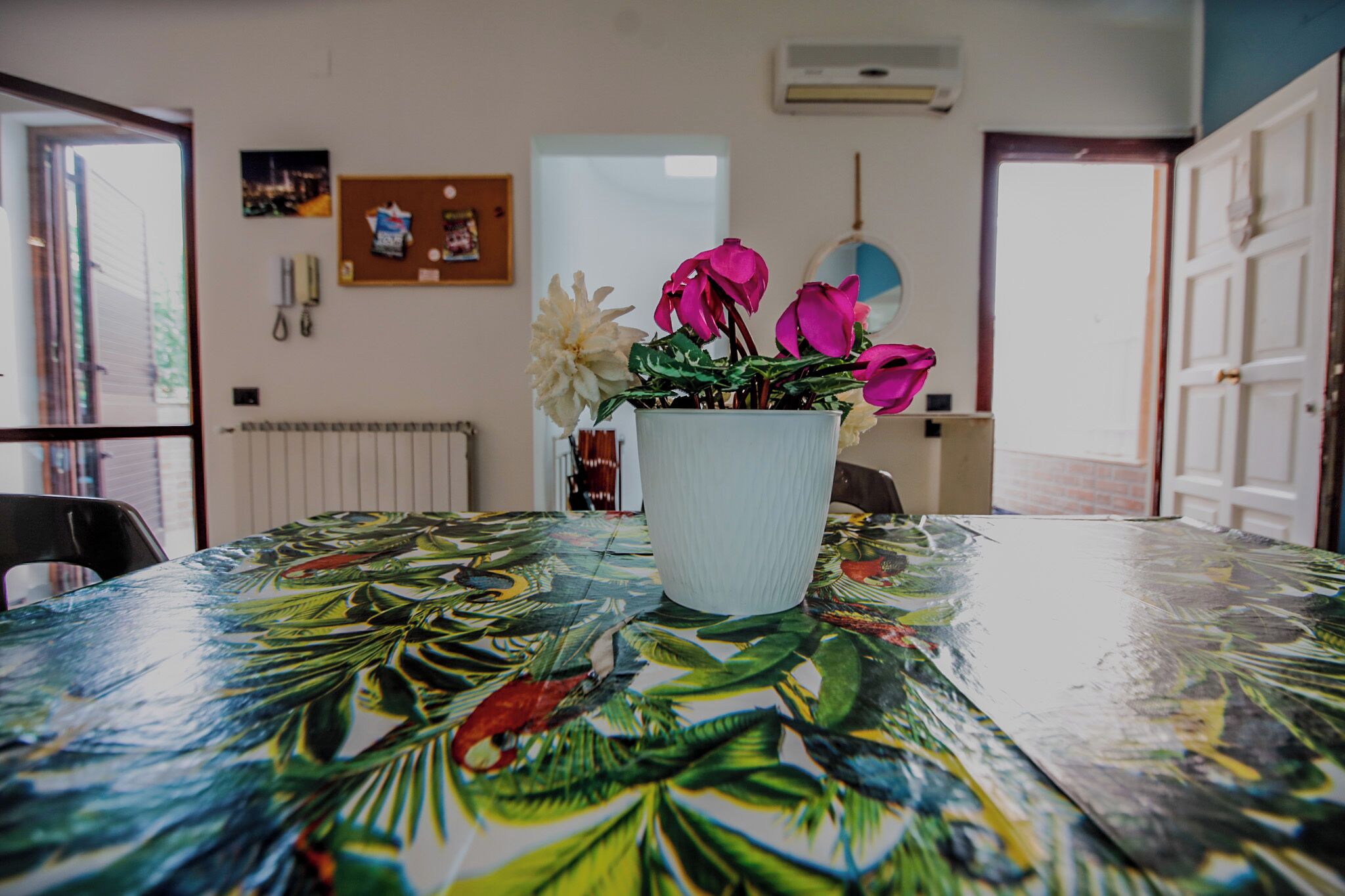Une table à manger ornée d’un motif de feuilles tropicales, un vase blanc contenant des fleurs roses et un climatiseur fixé au mur.