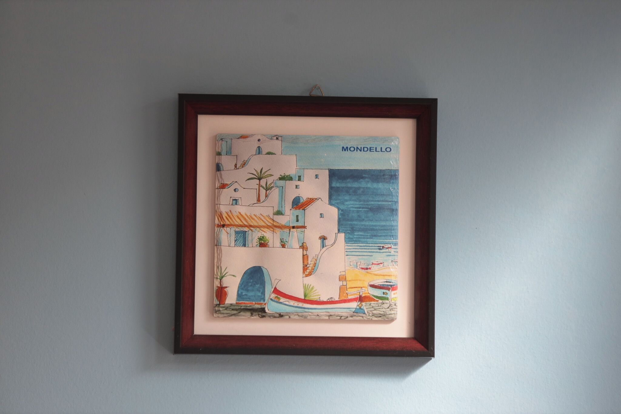 A framed artwork depicting a coastal scene with buildings, a boat, and the sea.