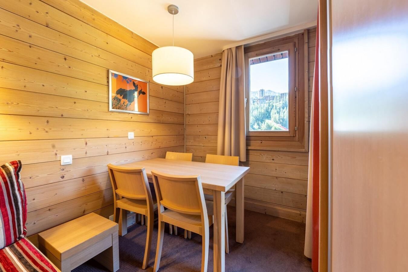 A wooden-paneled room with a dining table and chairs, a bench, a hanging light fixture, and a window with a view of a mountainous landscape.