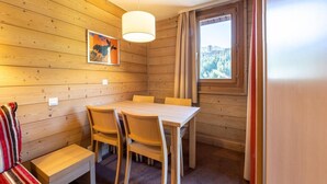 Une pièce aux murs lambrés, comprenant une table à manger et des chaises, un banc, un luminaire suspendu et une fenêtre donnant sur un paysage montagneux.