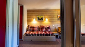 Une chambre avec une tête de lit en bois, un lit avec une literie à rayures, deux lampes fixées au mur et une petite table avec une chaise.