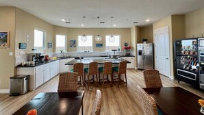 Una cocina moderna con una isla central, zona de comedor con sillas de mimbre y una máquina expendedora.