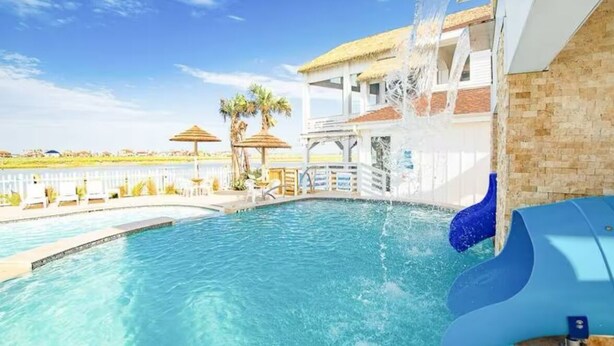 Una piscina con tobogán de agua, palmeras y un edificio con techo de paja al fondo.