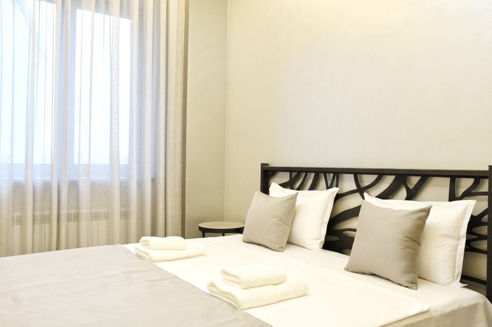 A neatly made bed with white linens and pillows, a round bedside table, and sheer curtains on a window.