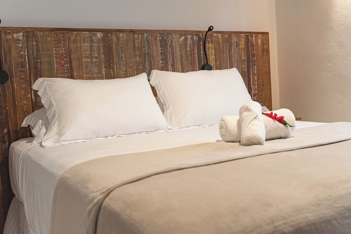 Un lit bien fait, avec du linge de lit blanc et une serviette décorative ornée d’une fleur rouge.