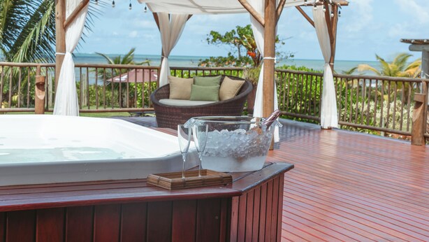 Une terrasse en bois avec un jacuzzi, une chaise en osier avec des coussins et un saladier en verre rempli de glace et d’une boisson.