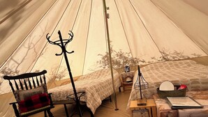 A tent interior with a bed, a chair, a coat rack, and a small table with a book and a lamp.