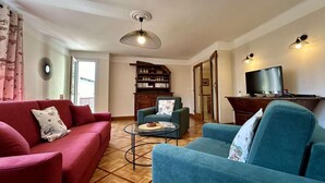 Una sala de estar con un sofá rojo, un sillón azul, una mesita redonda pequeña, una estantería y un televisor sobre un mueble de madera.