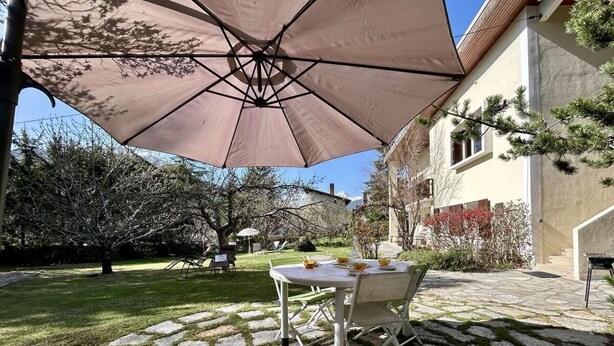 Un patio con una mesa y sillas bajo un paraguas grande, rodeado por un sendero de piedra y follaje.
