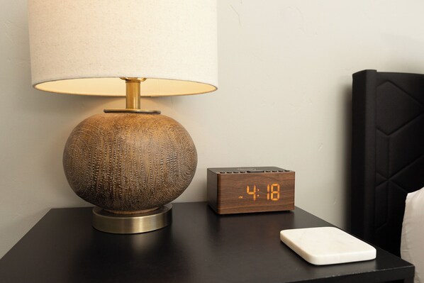 Een moderne lamp met een geribbelde voet en een cilindrische kap, een houten nachtkastje, een digitale klok waarop 4:18 staat, en een wit rechthoekig object op het tafeltje.