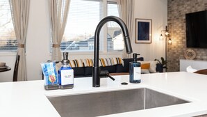A modern kitchen with a stainless steel sink, a black faucet, and dish soap. There is a window with curtains, a brick wall, and a flat-screen TV mounted above the counter. - Downtown Boise, BSU, Adventures! (Boise)