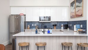 A modern kitchen with white cabinets, a stainless steel refrigerator, a microwave, and a gas stove. There are wooden stools at the island and a trash can to the left. - Downtown Boise, BSU, Adventures! (Boise)