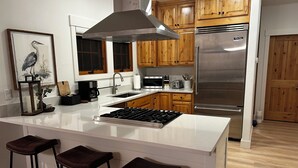 A modern kitchen with wooden cabinets, a stainless steel refrigerator, a gas stove, and three bar stools.