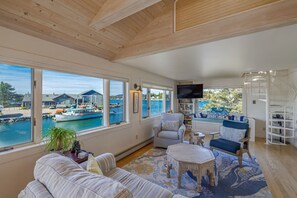Ein großzügiger Wohnraum mit großen Fenstern mit Blick auf eine Marina, eine gemütliche Sitzecke mit einer Couch und Sesseln sowie einem an der Wand befestigten Flachbildfernseher.