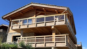 A two-story wooden cabin with a balcony on the upper floor, stone foundation, and a clear blue sky in the background.