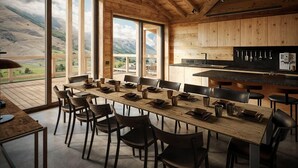 A dining area with a long wooden table set for 12, surrounded by chairs, with a kitchen area and large windows offering a mountain view. - Chalet, wifi, 300m from resort center, sauna, terrace, fireplace or stove, parking, tv, ski locker (Les Deux Alpes)
