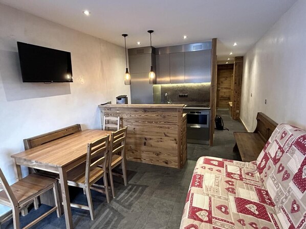 A compact living space with a wooden dining table and chairs, a sofa with a patterned cover, a kitchen area with a built-in island, and a flat-screen TV mounted on the wall. - 350m from shops, floor -2, view mountain, balcony, tv, 30m², Méribel-Mottaret (Les Allues)