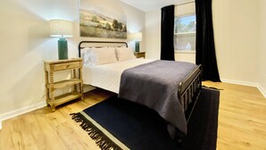 Ein Schlafzimmer mit Bett, Nachttisch, Lampe, Deckenventilator, einem Fenster mit Vorhängen und einem Bild an der Wand.