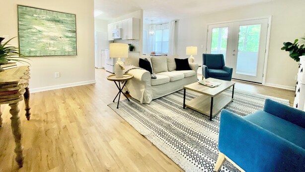Ein modernes Wohnzimmer mit einer Couch, einem Couchtisch und einem blauen Sessel. Im Hintergrund befindet sich eine Küche mit weißen Schränken und einer Mikrowelle.