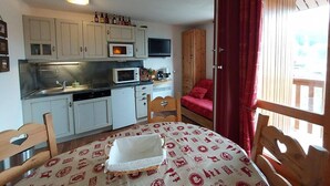 A compact kitchen with white cabinets, a microwave, and a coffee maker. There is a dining table with a white tablecloth and a red patterned table runner. A wooden chair with a heart-shaped cutout is visible.