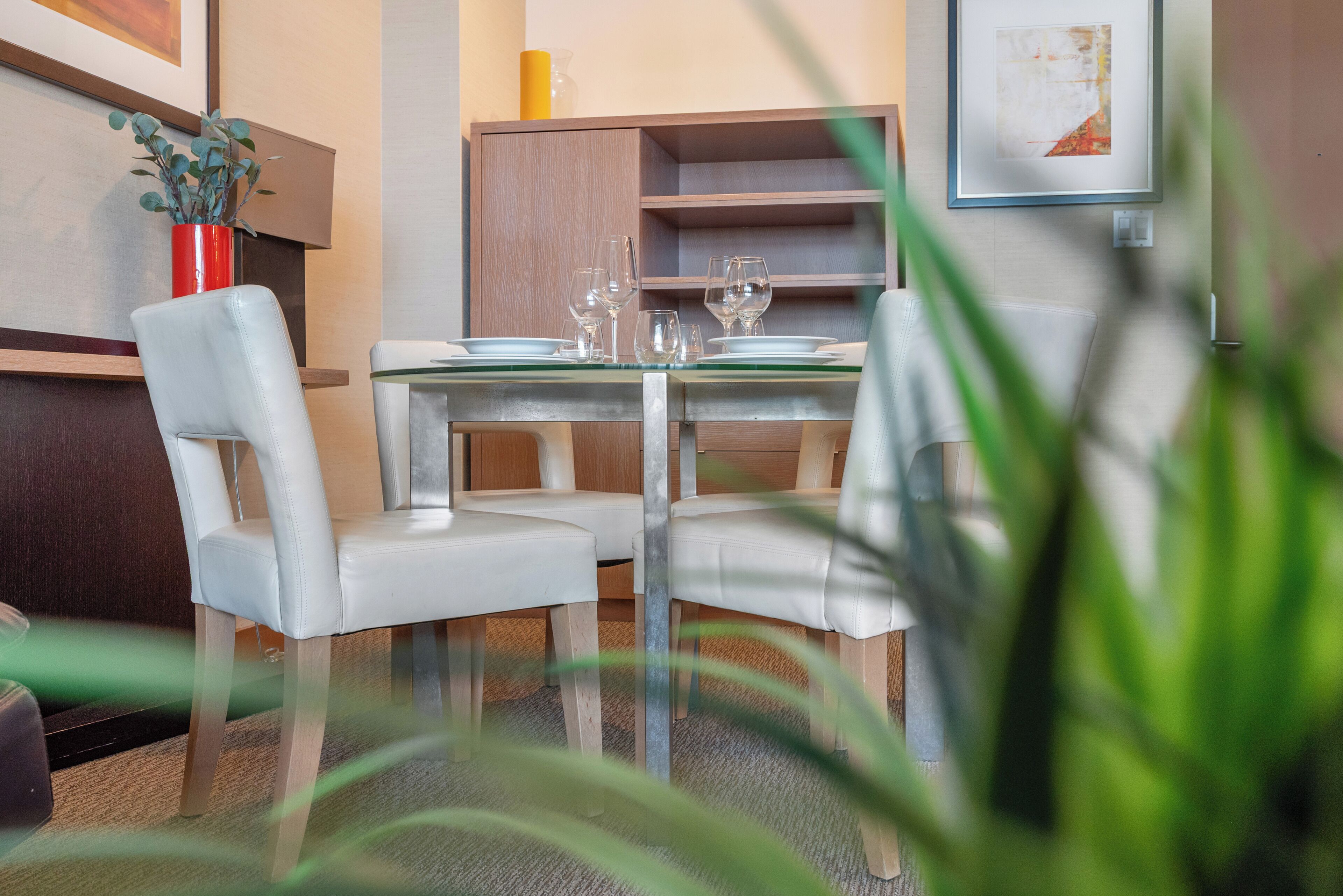 A dining area with a glass table, white chairs, and a wooden cabinet in the background.
