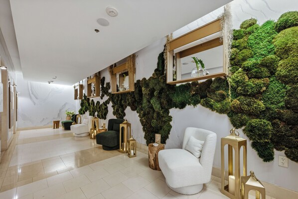 Un interior moderno con un sofá blanco, un sillón oscuro y una silla blanca. Hay dos espejos con marco dorado en la pared, una mesita de madera y una pequeña planta en maceta.