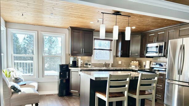 A modern kitchen with dark wood cabinets, stainless steel appliances, and a large island with bar stools. - Cozy Home on Sturgeon Lake with Hot Tub (Kawartha Lakes)