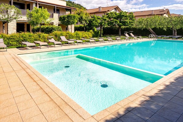 Una piscina rectangular con agua azul cristalina, rodeada por un solárium empedrado. Hay sillones de descanso a lo largo de la piscina.