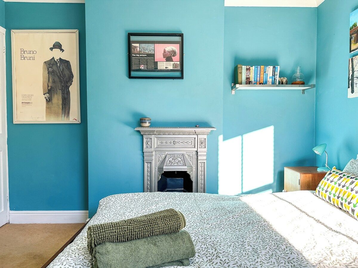 Un dormitorio con cama, chimenea, estante con libros y un cuadro en la pared.