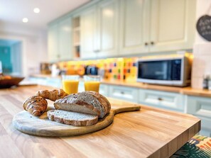 A breakfast setting with bread, croissants, and orange juice on a wooden table in a kitchen with light-colored cabinets and a microwave. - Charming Cottage Near Cathedral w/ Gated Parking (Canterbury)