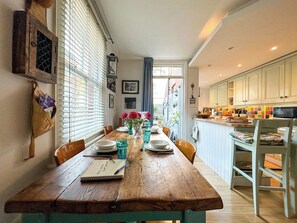 A dining area with a wooden table set for four, a kitchen with white cabinets, and a window with blinds. - Charming Cottage Near Cathedral w/ Gated Parking (Canterbury)
