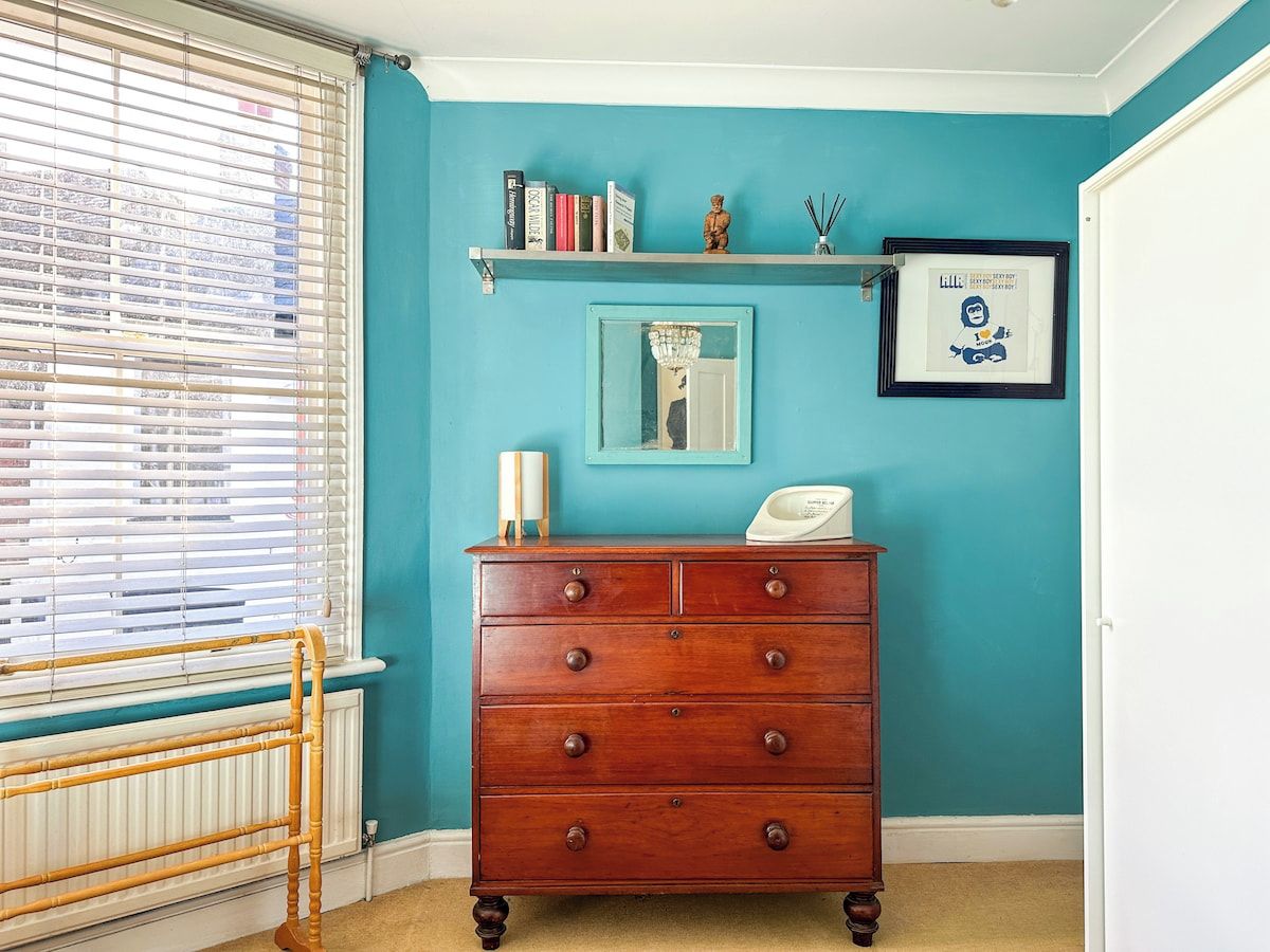 A room with teal walls, a wooden dresser with drawers, a shelf with books and a small statue, a framed picture, and a window with blinds.
