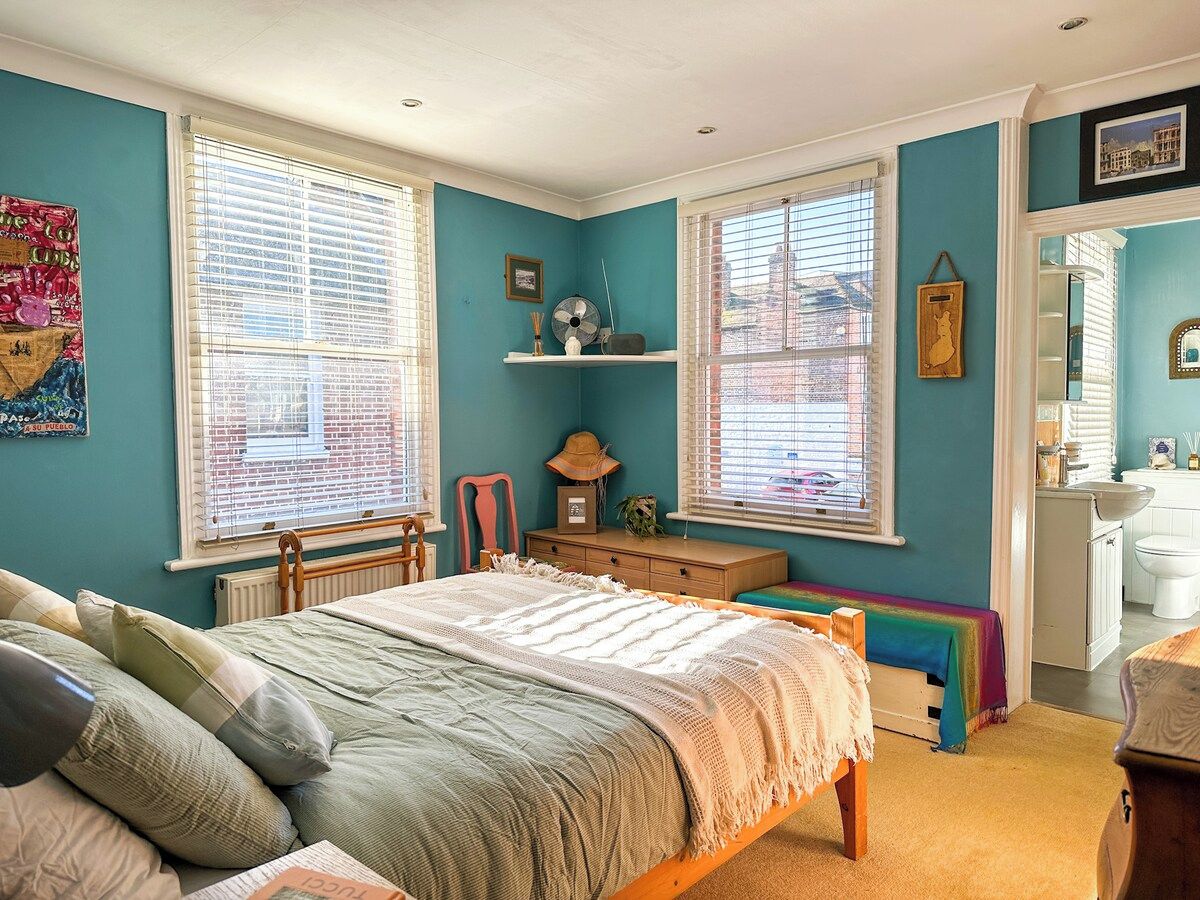 Une chambre à coucher avec un lit, un bureau et une chaise. On y trouve des fenêtres avec des stores, un radiateur et une étagère ornée d’objets décoratifs.