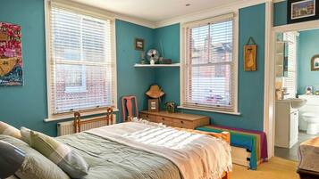 Une chambre à coucher avec un lit, un bureau et une chaise. On y trouve des fenêtres avec des stores, un radiateur et une étagère ornée d’objets décoratifs.