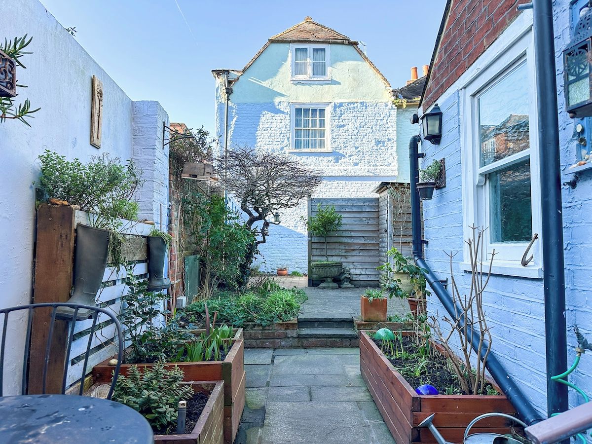 Un étroit sentier de jardin mène à une maison dont la façade est en pierre et les murs extérieurs peints en bleu.