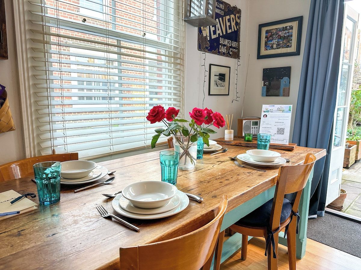 Une table à manger dressée avec des assiettes, des couverts et des verres bleus, et un vase rempli de fleurs rouges placé au centre.