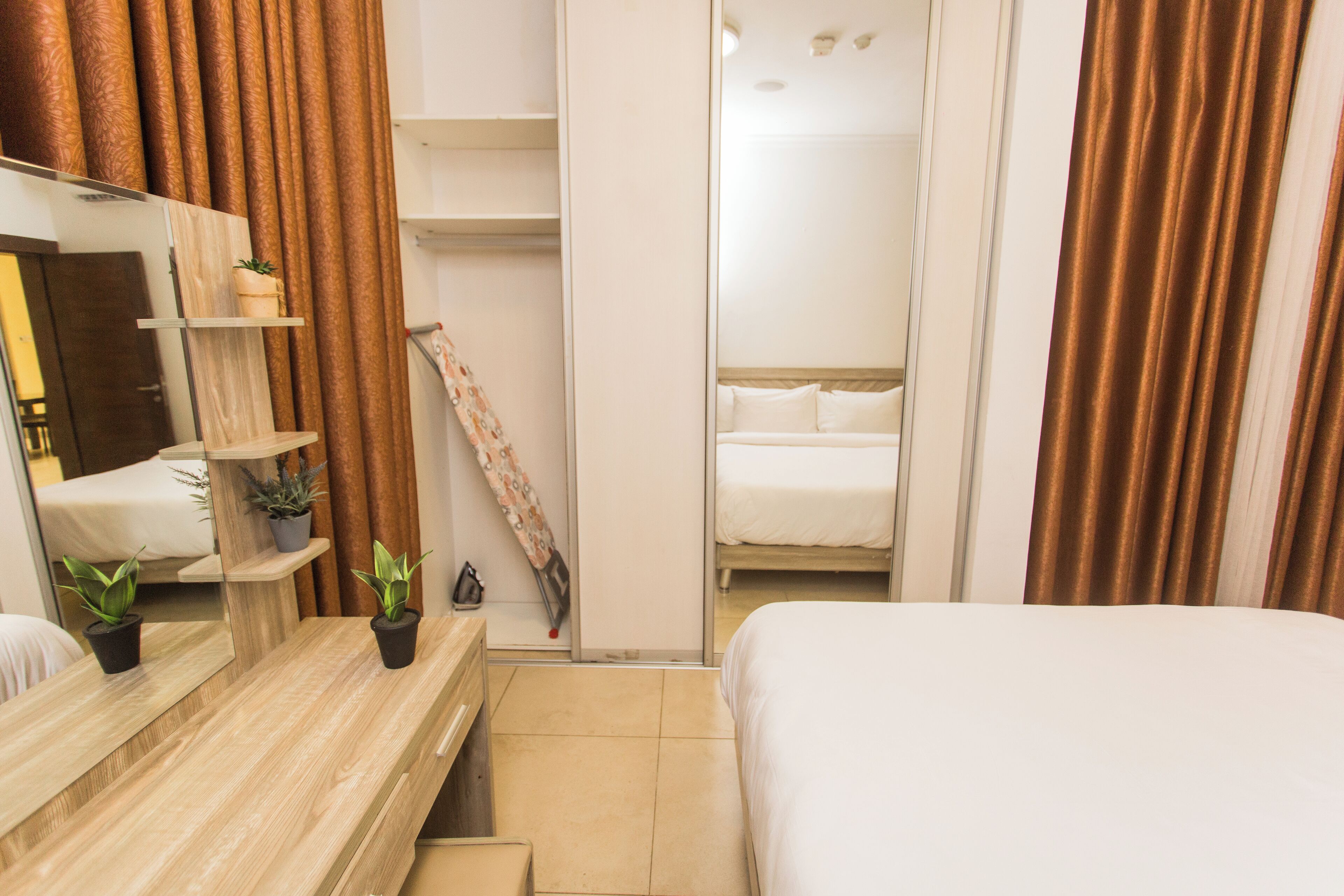 A hotel room with a wooden vanity, a bed, a mirror, a shelf with potted plants, and a sliding door leading to a bathroom.