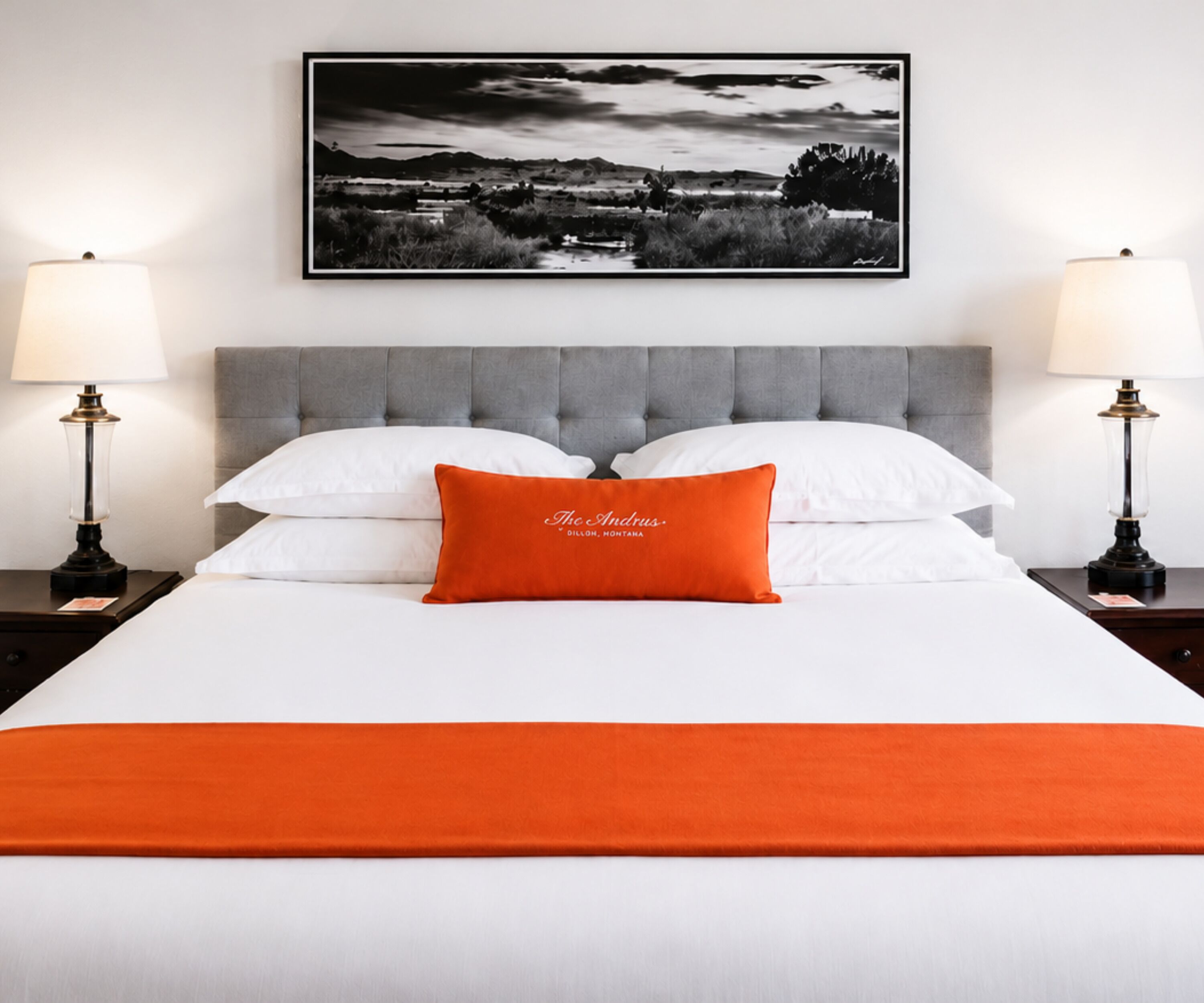 A neatly made bed with white linens and a single orange pillow, flanked by two bedside tables with lamps, and a framed black and white landscape picture above.