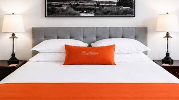 A neatly made bed with white linens and a single orange pillow, flanked by two bedside tables with lamps, and a framed black and white landscape picture above.