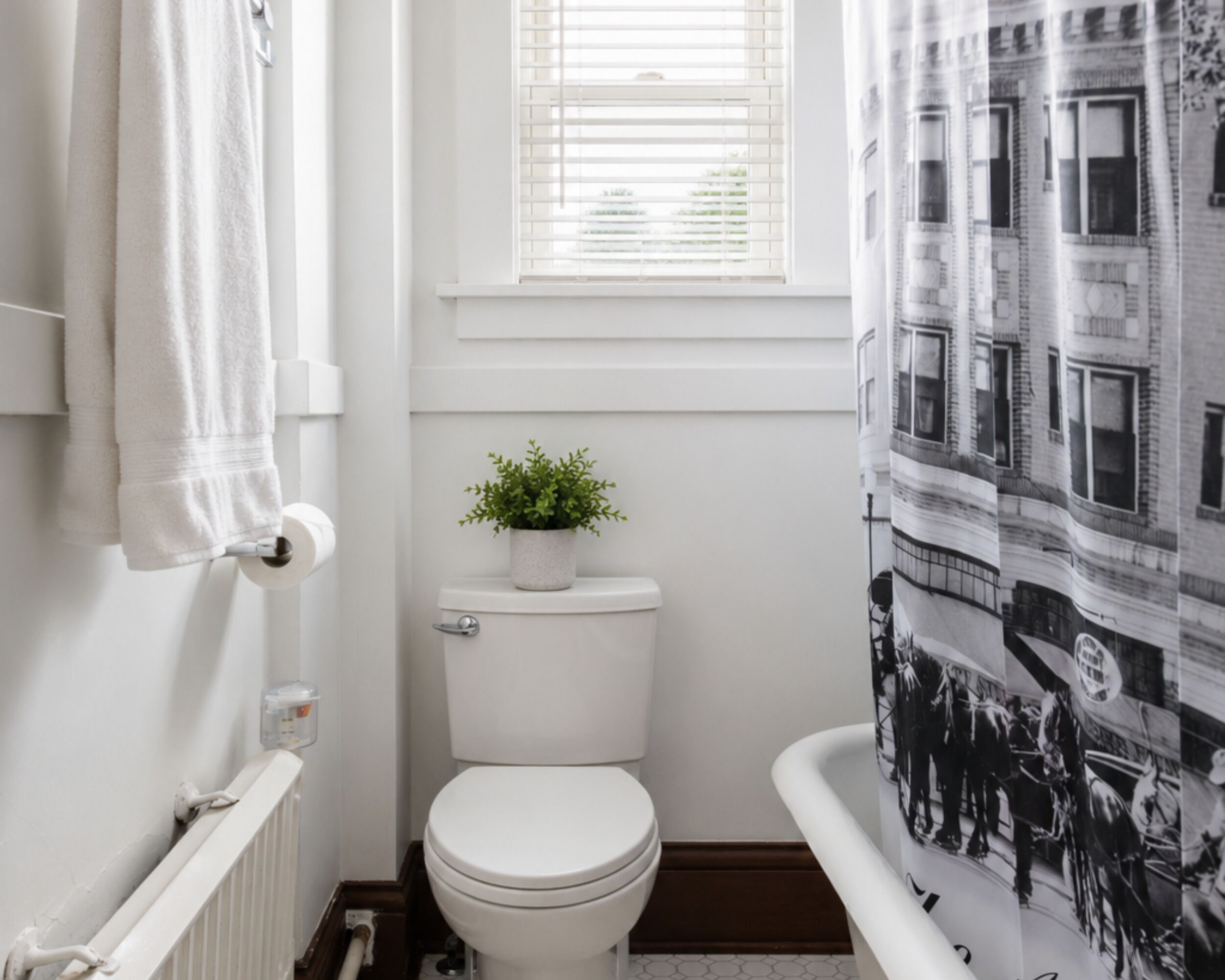 A bathroom with a toilet, a window with blinds, a towel rack, and a shower curtain with a black and white print.
